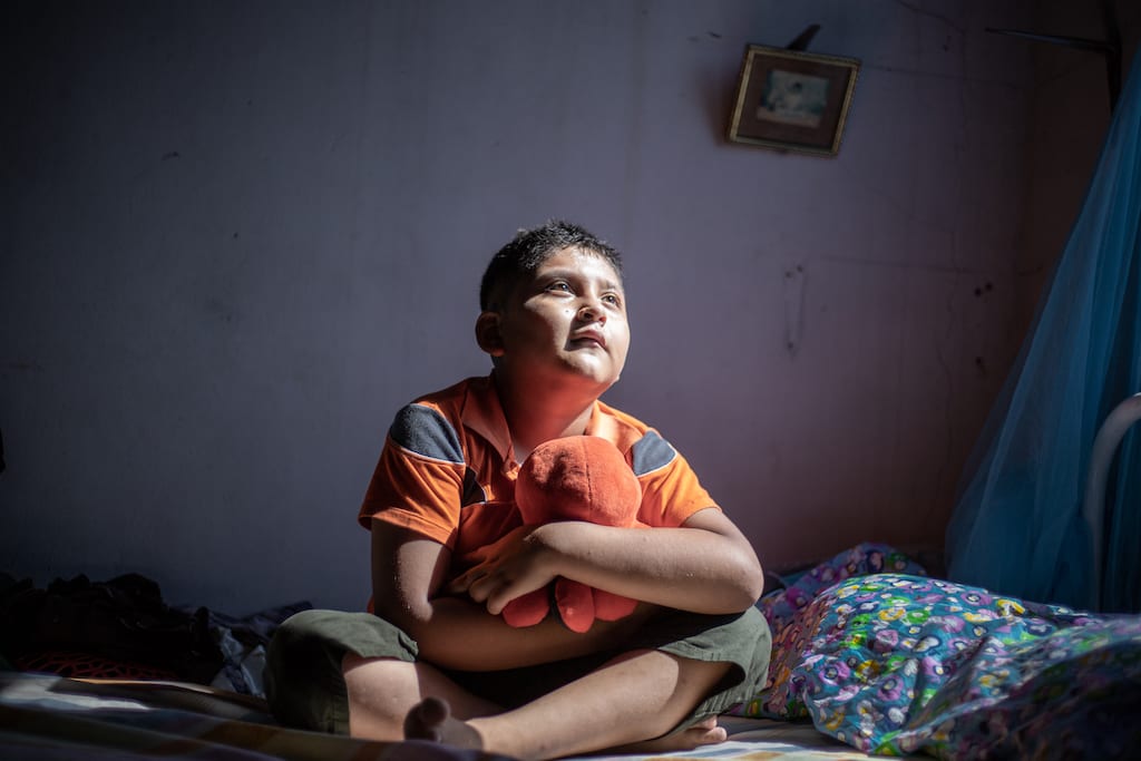 Boy from Ecuador sits cross-legged on his bed holding a stuffed animal.